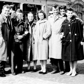 Group of people posing outside a train station in mid-20th century attire.