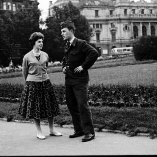 Two people standing and conversing outdoors in 1950s-style attire.