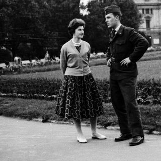 Two people standing and conversing outdoors in 1950s-style attire.
