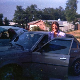 Woman smiling from car roof, suburban driveway.