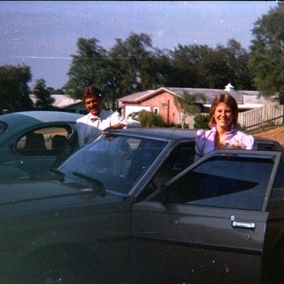 Two people posing by cars in a residential driveway.