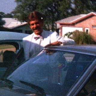 Two people posing by cars in a residential driveway.