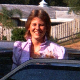 Two people posing by cars in a residential driveway.
