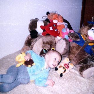 Baby lies on a rug surrounded by stuffed animals.