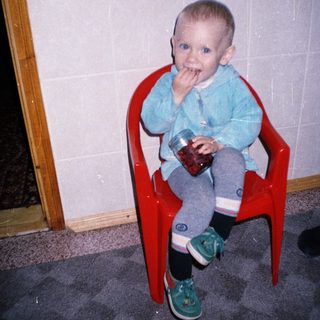 Here's a brief summary of the image: Toddler sits in a chair holding a jar, eating.