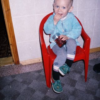 Here's a brief summary of the image: Toddler sits in a chair holding a jar, eating.