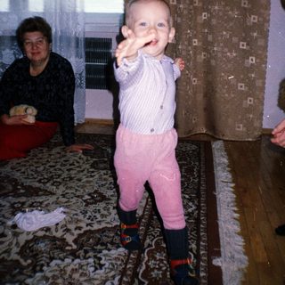 Baby in patterned clothing stands, with adults nearby.