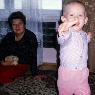 Baby in patterned clothing stands, with adults nearby.