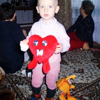 Toddler holds a heart-shaped toy, with adults in background.