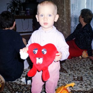 Toddler holds a heart-shaped toy, with adults in background.
