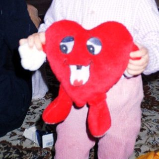Toddler holds a heart-shaped toy, with adults in background.