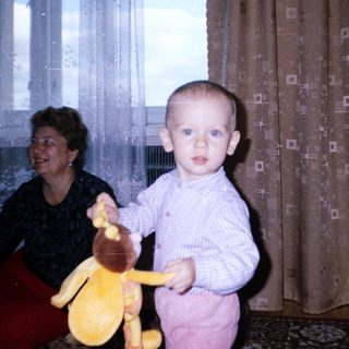 Baby holding toy, standing near a woman and curtains.