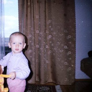 Baby holding toy, standing near a woman and curtains.