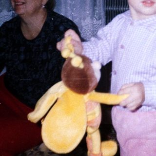 Baby holding toy, standing near a woman and curtains.