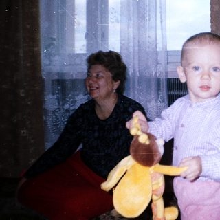 Baby holding toy, standing near a woman and curtains.