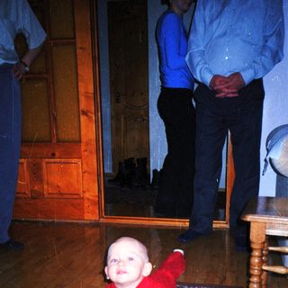 Baby crawls on a rug with a man in the background.