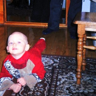 Baby crawls on a rug with a man in the background.