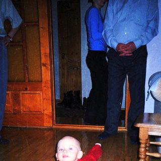 Baby crawls on a rug with a man in the background.