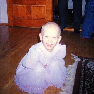 Here's a brief summary of the image: Baby in a dress sits on a rug.