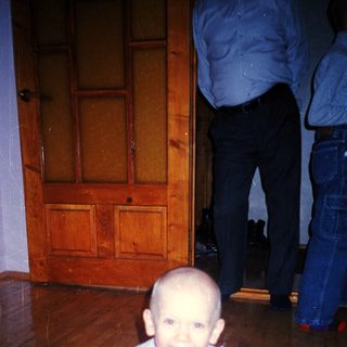 Here's a brief summary of the image: Baby in a dress sits on a rug.