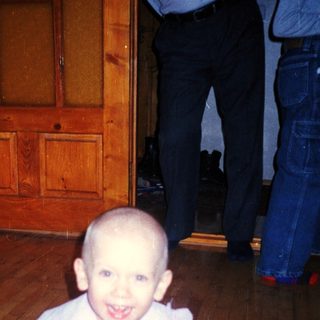 Here's a brief summary of the image: Baby in a dress sits on a rug.