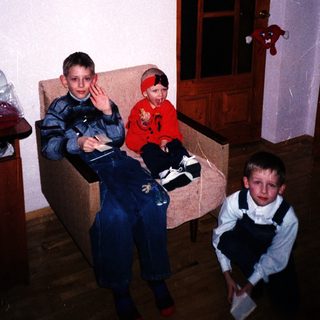 Three boys in overalls sit and stand in a room.