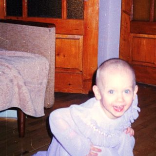 Here's a brief summary of the image: Young child with a shaved head kneels near a door.