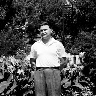 Man stands in a garden, smiling, surrounded by foliage.