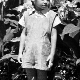 Here's a brief summary of the image: Young boy stands near foliage, with a house in background.