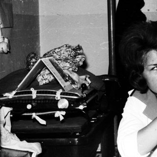 Here's a brief summary of the image: Woman sits in a sparse room, looking directly at the camera.