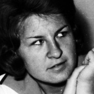 Here's a brief summary of the image: Woman sits in a sparse room, looking directly at the camera.