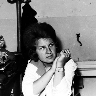 Here's a brief summary of the image: Woman sits in a sparse room, looking directly at the camera.