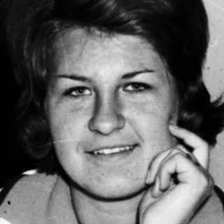 Woman sits near a sink and trunk, looking at camera.