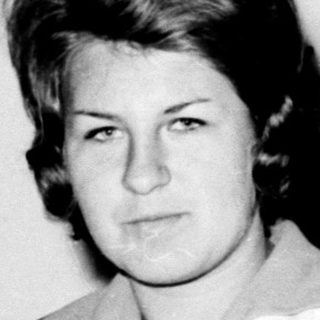 Black and white portrait of a woman standing near a coat rack.