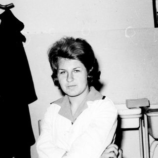 Black and white portrait of a woman standing near a coat rack.