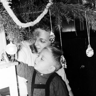 Boy in overalls sits near a decorated Christmas tree.
