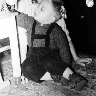 Boy in overalls sits near a decorated Christmas tree.