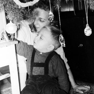 Boy in overalls sits near a decorated Christmas tree.