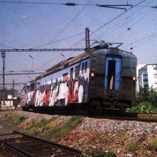 Graffiti-covered train on tracks, with buildings and power lines.