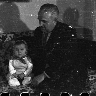 Here's a brief summary of the image: Baby sits on a couch with an adult nearby.