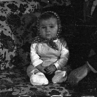 Here's a brief summary of the image: Baby sits on a couch with an adult nearby.