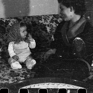 Baby sits on a couch with a woman nearby.