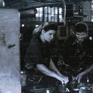 Two workers operate machinery in what appears to be a factory.