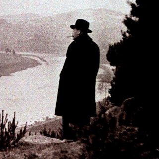 Man in coat stands overlooking a river and landscape.