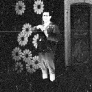Woman stands near a building with flowers and an archway.