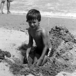 Boy sits in a hole on a beach, woman in background.
