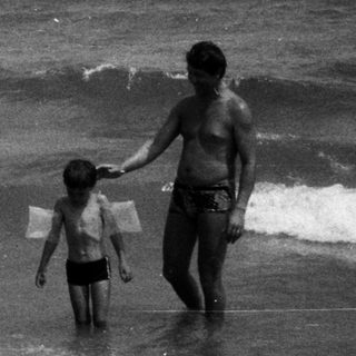 Man and child walk in ocean waves on a beach.