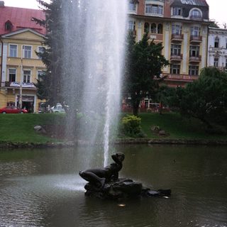 Fountain with statue in a park, buildings in background.