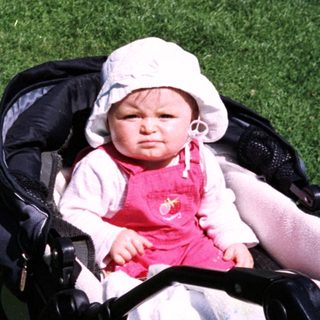 Baby in stroller sits on grass, wearing a hat.