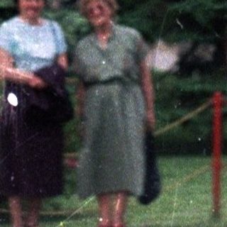 Two women stand near a building with evergreen trees.